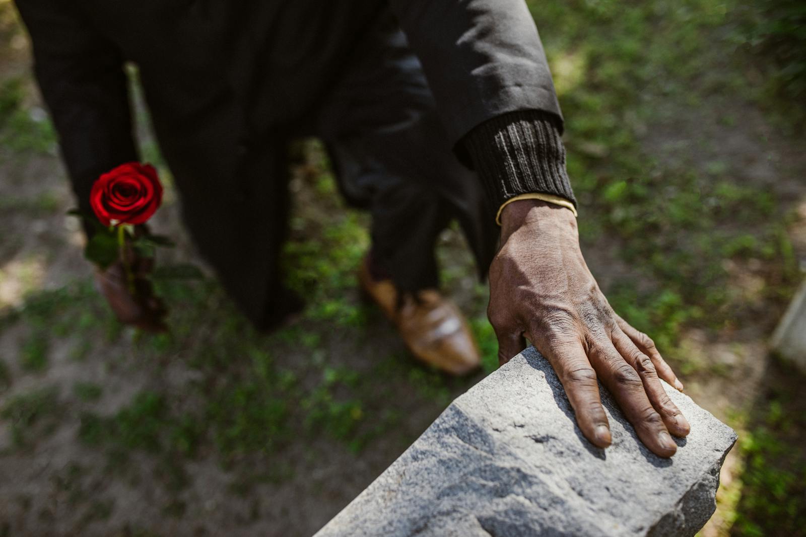 A man mourns a loved one, holding a rose by a gravestone, signifying loss and remembrance.